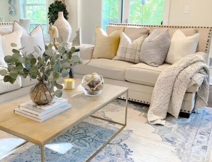 Un salon lumineux, ambiance douce et naturelle : canapé beige en lin, plaid en laine posé négligemment, tapis berbère au sol, quelques coussins texturés, une plante verte en coin, lumière chaude en fin de journée et une bougie allumée sur une table basse en bois brut.