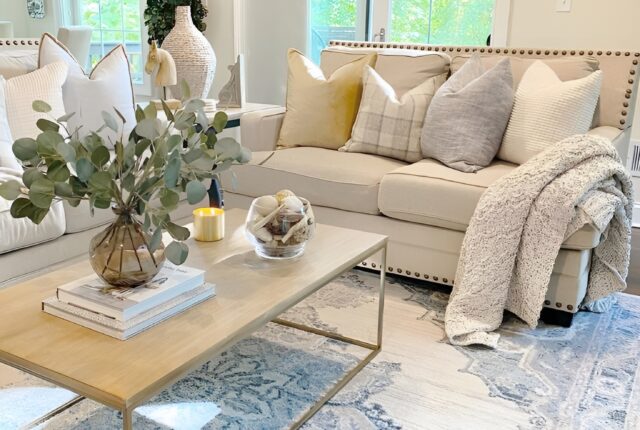 Un salon lumineux, ambiance douce et naturelle : canapé beige en lin, plaid en laine posé négligemment, tapis berbère au sol, quelques coussins texturés, une plante verte en coin, lumière chaude en fin de journée et une bougie allumée sur une table basse en bois brut.