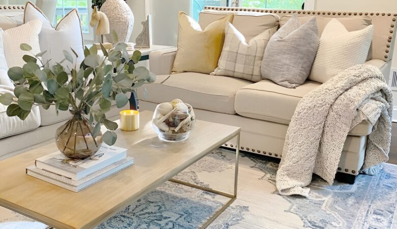 Un salon lumineux, ambiance douce et naturelle : canapé beige en lin, plaid en laine posé négligemment, tapis berbère au sol, quelques coussins texturés, une plante verte en coin, lumière chaude en fin de journée et une bougie allumée sur une table basse en bois brut.