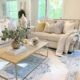 Un salon lumineux, ambiance douce et naturelle : canapé beige en lin, plaid en laine posé négligemment, tapis berbère au sol, quelques coussins texturés, une plante verte en coin, lumière chaude en fin de journée et une bougie allumée sur une table basse en bois brut.