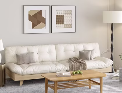 Un salon baigné de lumière naturelle, canapé en lin beige avec coussins doux, plaid en laine, table basse ronde en bois clair, lampe sur pied minimaliste, tapis structuré, et une plante haute dans un pot blanc mat.