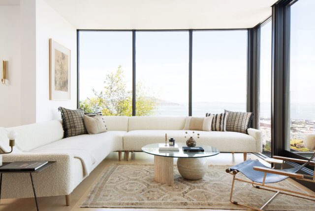 un salon lumineux avec de grandes baies vitrées, un canapé aux lignes droites en beige clair, une table basse en bois brut, et une plante verte