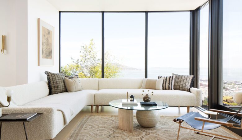 un salon lumineux avec de grandes baies vitrées, un canapé aux lignes droites en beige clair, une table basse en bois brut, et une plante verte