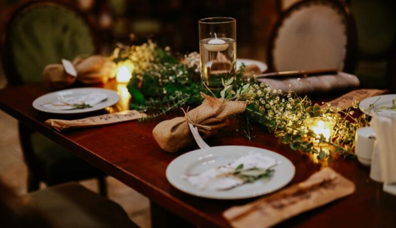 une table de Noël vue de face avec nappe en lin beige, bougies blanches, branches de sapin, petites décorations dorées et mini cadeaux sur chaque assiette